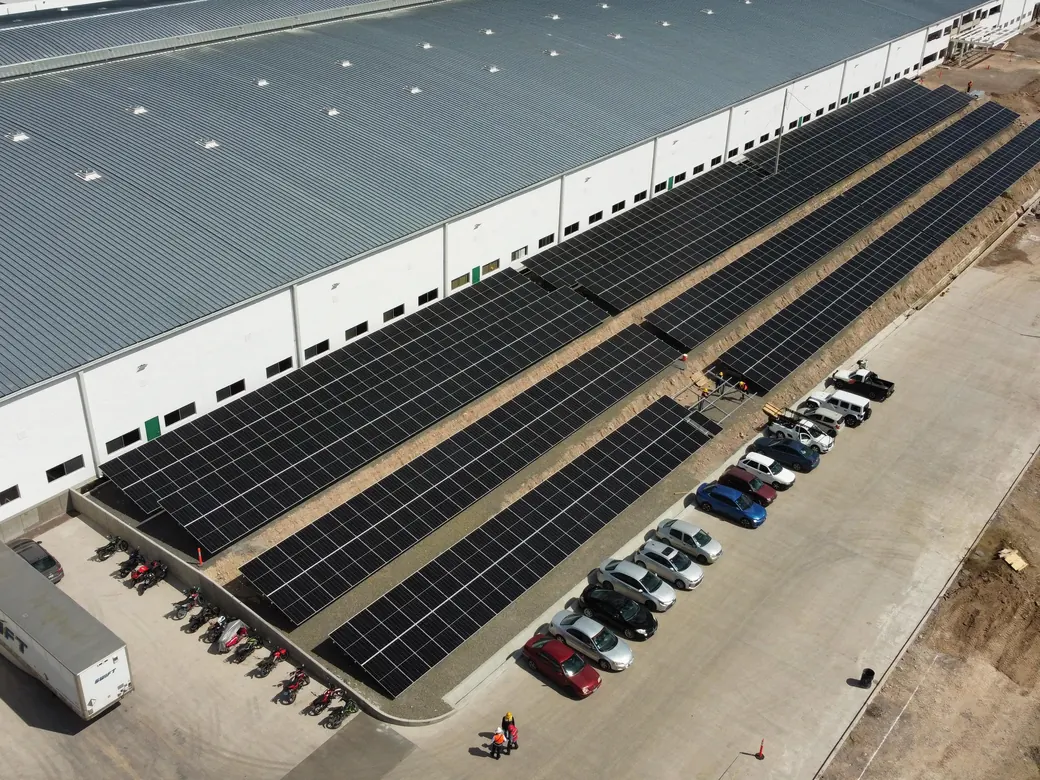 Hileras de módulos solares hincadas al suelo a lo largo de una nave industrial nueva, junto al estacionamiento